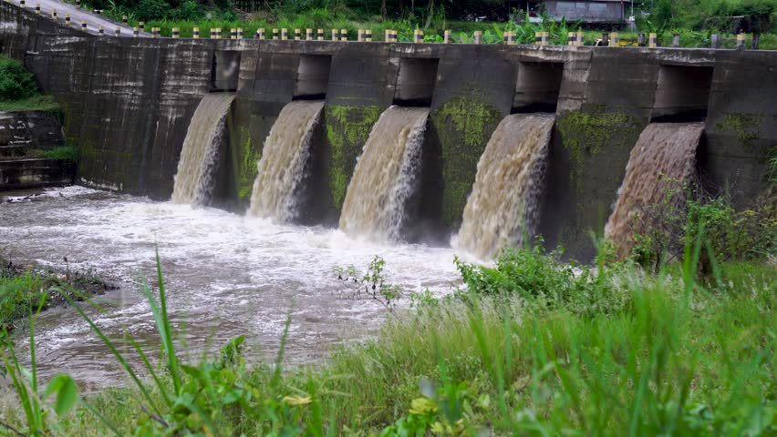 This slowly panning video from right to left shows a large concrete dam or floodgate structure. Abundant, murky river water rushes through five main channels simultaneously.