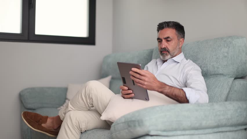Focused middle aged man comfortably relaxing on the couch in his living room. Handsome person browsing the internet or reading on a tablet