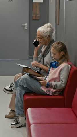 Vertical shot of young child in clinic waiting room using tablet while sitting on couch with grandma speaking by smartphone