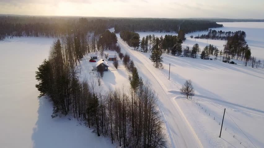 Aerial view of Scenic Winter Landscape Featuring a Remote Cabin Surrounded by Snow-Covered Fields and Tall Trees in a Serene, Sunlit Atmosphere in Finland