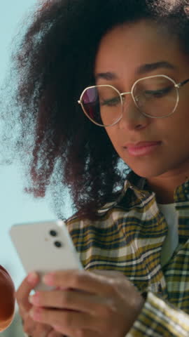 Two African American students sit using mobile phones scrolling online outdoors ethnic friends woman girl female man male guy together gadget addict smartphones social media in city college university