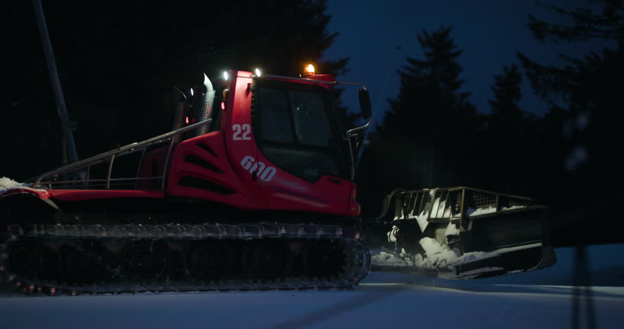 Red snowcat with bright lights on a snowy ski slope at night. A chairlift and snow-covered trees are visible in the background.
