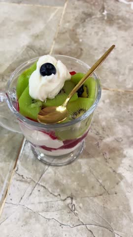 Vertical 4K 60fps video of homemade fruit salad parfait with yogurt, fresh kiwi, raspberries, and blueberries. Healthy breakfast dessert in a glass cup with a gold spoon on a marble table.