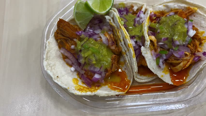Three authentic Cochinita Pibil tacos with green salsa, chopped red onions, and lime wedges in a plastic takeout container. Delicious Mexican street food lunch served on a wooden table.