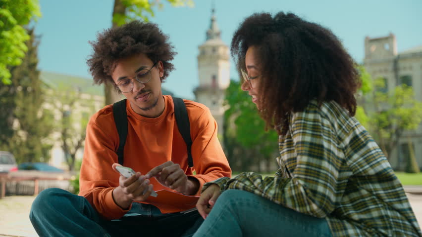 African American students sitting together looking at mobile phone discussing ethnic people classmates woman girl female man male guy outside in city talking smartphone university college high school
