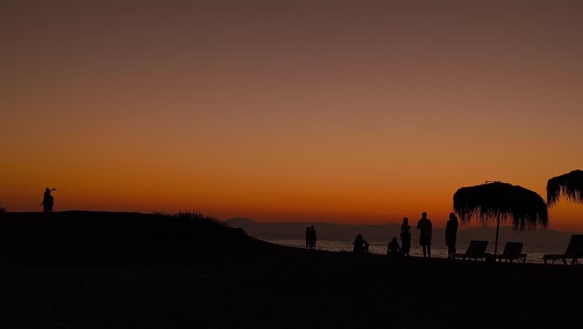 People and beach umbrellas made of palm leaves in the warm orange light of a sea sunset, silhouettes against the sky and the sea.