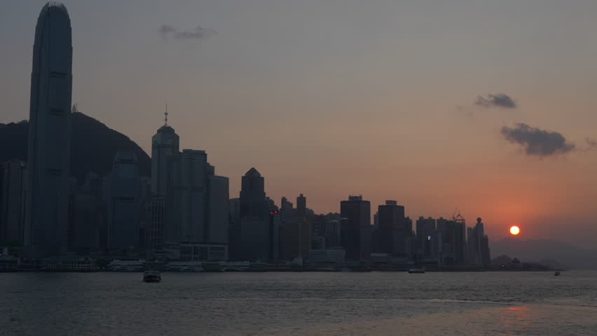 Dusk Over Hong Kong’s Victoria Harbour: Skyscrapers Frame a Warm Orange Sky, Sunset Blushes Clouds Over Hills, Boat Shadows Ripple Through Golden Sunlight.
