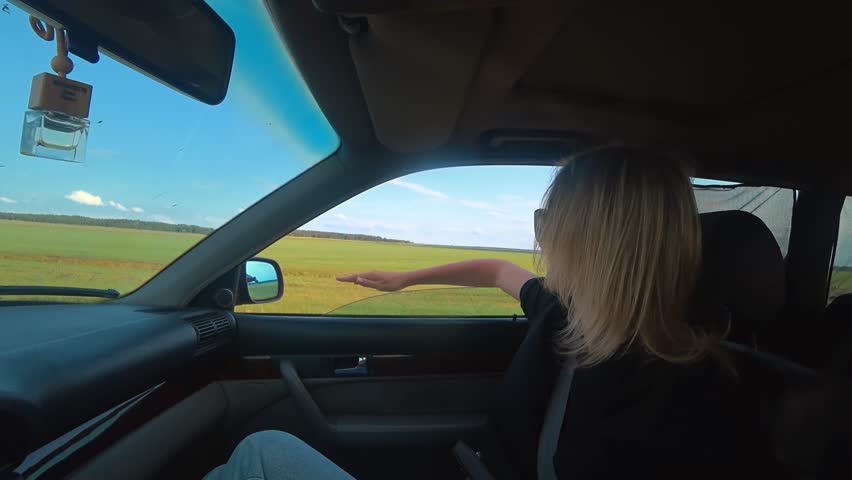 Joyful blonde woman sitting in the passenger seat of a moving car, sticking her hand out the open window to feel the air and enjoying the freedom of a scenic countryside road trip