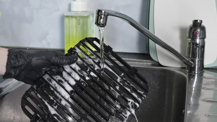 Close-up of a gloved hand meticulously cleaning a black air fryer griddle under running water with suds in a stainless steel kitchen sink, highlighting routine household cleaning