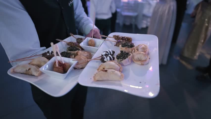 Slow‑motion shot tracks a waiter in a white shirt and dark vest as he carries two large white trays filled with bite‑size appetizers and dipping sauces, moving through a softly lit reception hall