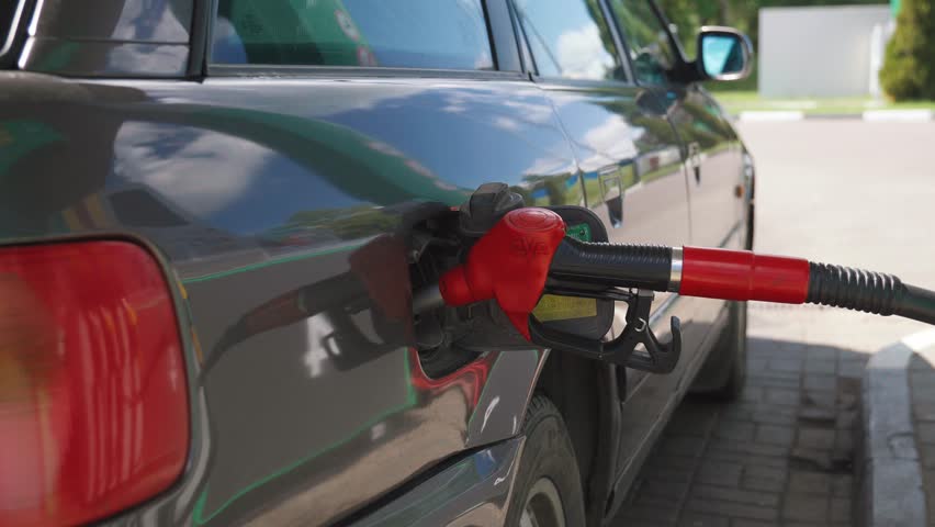 Red fuel nozzle inserted into a dark gray car's tank at a gas station for refueling, representing rising gasoline prices, energy crisis, and the cost of transportation and travel