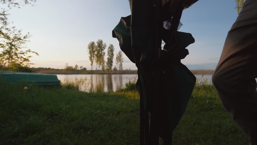 Man setting up folding chair on grassy lakeshore during beautiful sunset, preparing for relaxing evening of fishing or camping in quiet nature