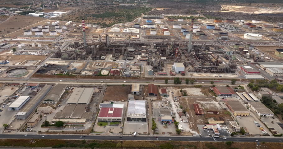 Aerial view of a large, complex oil and chemical refinery. The facility features a tall  chimney, dense industrial piping and machinery, and a vast storage area of cylindrical tanks.