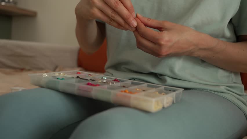 Imaginative woman threading colorful beads onto string to create handmade bracelets and necklaces, seated at home with a bead organizer box on her lap, focused on detailed jewelry making
