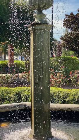 Backlit droplets fall from a stone fountain beside trimmed hedges and greenery. A child wearing a helmet rides past, adding motion and play to the sunny park scene.