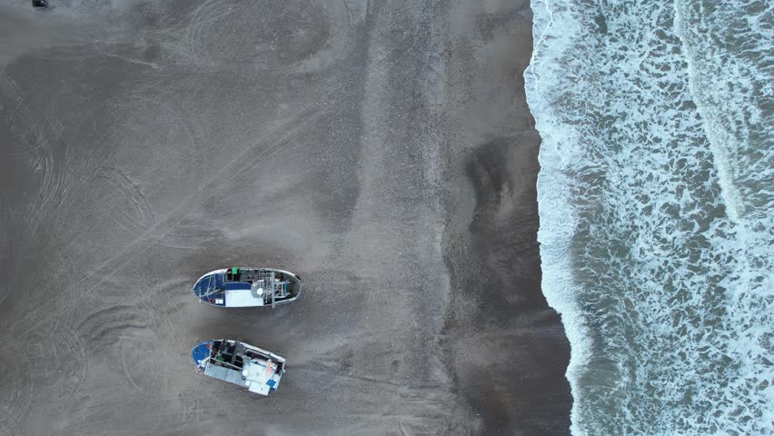 Aerial view of fishing boats pulled ashore on the Danish west coast, waves rolling in under heavy clouds at dusk.
