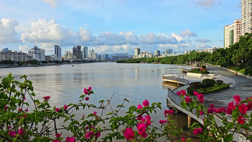 Scenic urban riverfront in Sanya with modern skyline, curved waterfront promenade, and pink flowers. Beautiful city landscape featuring calm water, pedestrian walkway, green trees, and cloudy blue sky