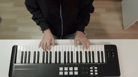 Top view of musician's hands playing black and white MIDI keyboard synthesizer with control panel buttons and knobs on white desk, creating music in home studio setup. - Powered by Shutterstock - Get 15% off with code: PIKWIZARD15