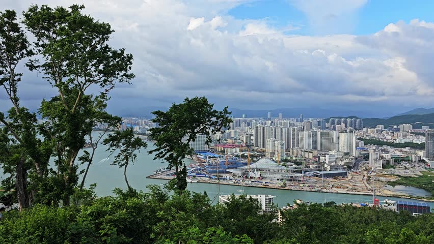 Scenic panoramic view of Sanya from Deer Turned Head Park, featuring a modern skyline, marina, mountains, lush greenery, and waterfront under a bright tropical sky.