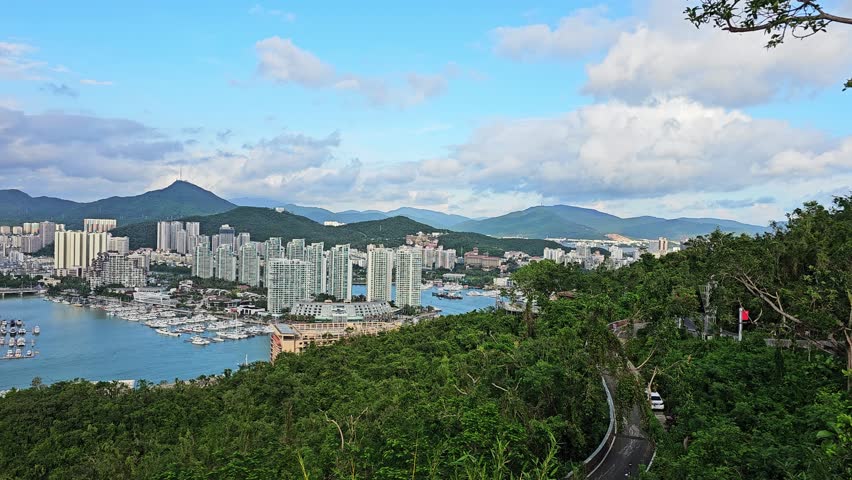 Scenic panoramic view of Sanya from Deer Turned Head Park, featuring a modern skyline, marina, mountains, lush greenery, and waterfront under a bright tropical sky.