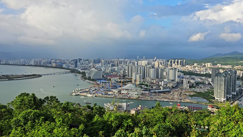 Scenic panoramic view of Sanya from Deer Turned Head Park, featuring a modern skyline, marina, mountains, lush greenery, and waterfront under a bright tropical sky.