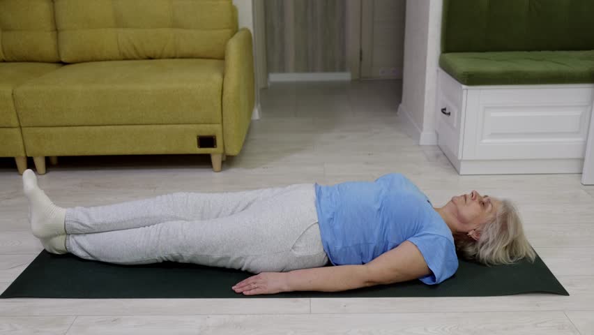 Elderly woman in blue shirt lying on dark green exercise mat performing leg lift movement. Core strengthening workout focusing on abdominal muscles in modern living room.