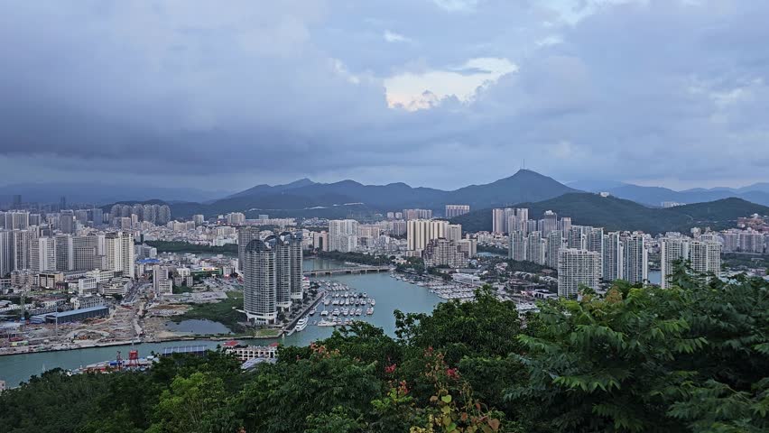 Scenic panoramic view of Sanya from Deer Turned Head Park, featuring a modern skyline, marina, mountains, lush greenery, and waterfront under a bright tropical sky.