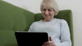 Senior woman in light gray cable-knit sweater holding tablet with both hands on green couch. Engaged digital reading showing technology adoption and mobile device use at home. - Powered by Shutterstock - Get 15% off with code: PIKWIZARD15