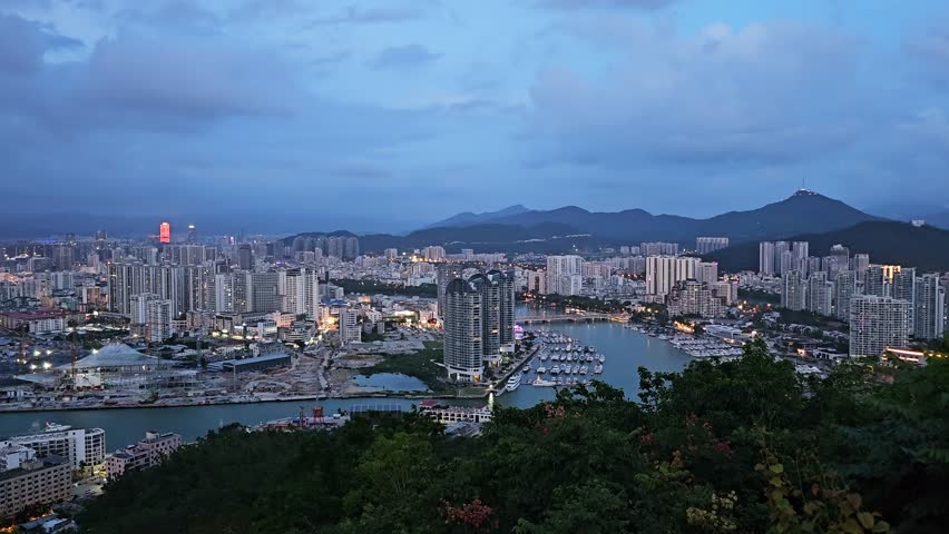 Scenic panoramic view of Sanya from Deer Turned Head Park, featuring a modern skyline, marina, mountains, lush greenery, and waterfront under a bright tropical sky.