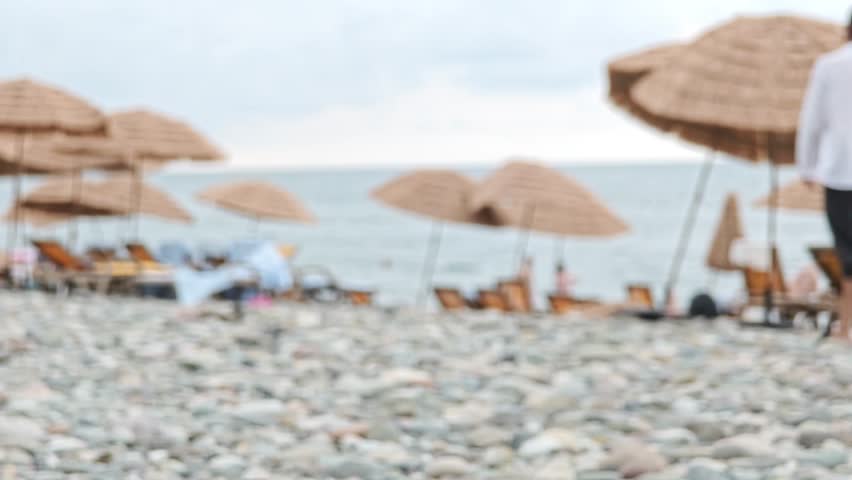 Indistinct background capturing a man strolling towards the ocean along a pebbled shoreline. Beachgoers lounging under straw umbrellas on a overcast summer day