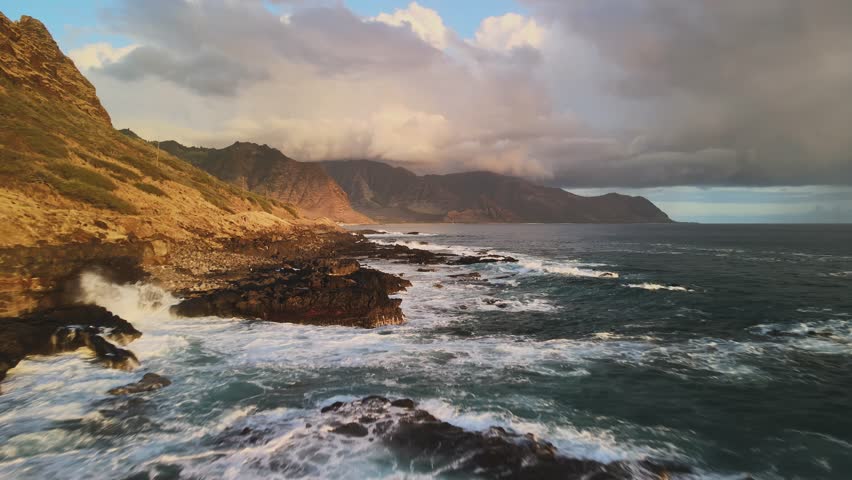 Kaena Point State Park. The rugged coastline of Hawaii with waves hitting the rocks. The sun sets behind mountains, casting warm light on the water and land.
