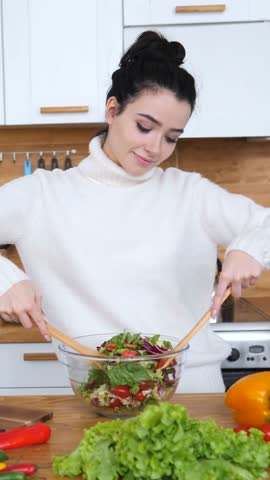 Attractive smiling woman in a cozy kitchen preparing a healthy meal, mixing fresh green lettuce, tomatoes, and other vegetables in a glass bowl with wooden spoons for a nutritious diet