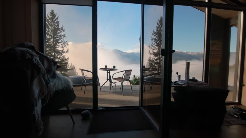 View through the open glass door onto a wooden terrace with table and chairs, looking at the amazing foggy mountain landscape in the morning