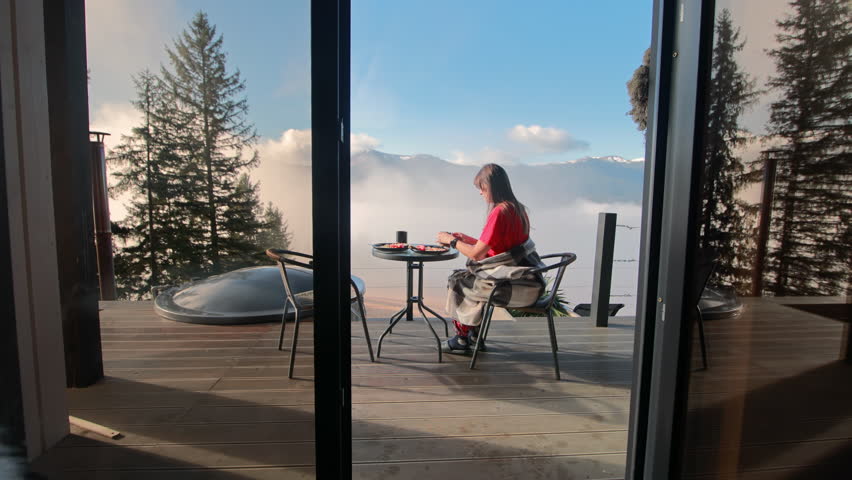 Young woman sitting on the terrace and drinking coffee in the morning, enjoying beautiful view of the foggy mountains through the glass door