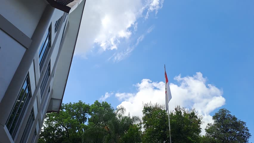 Handheld of video Indonesian flag waving on a tall pole above a government building under a bright clear sky, capturing patriotic spirit, national pride, and peaceful atmosphere. Blue sky, cloudy.