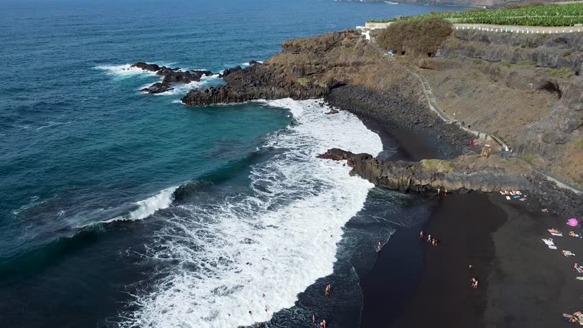 Drone aerial view of Playa El Bollullo, a stunning black sand beach in Tenerife with waves crashing along the shore, surrounded by rocky cliffs and sunbathing visitors	