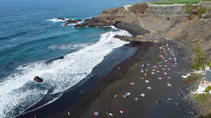 Drone aerial view of Playa El Bollullo, a stunning black sand beach in Tenerife with waves crashing along the shore, surrounded by rocky cliffs and sunbathing visitors	