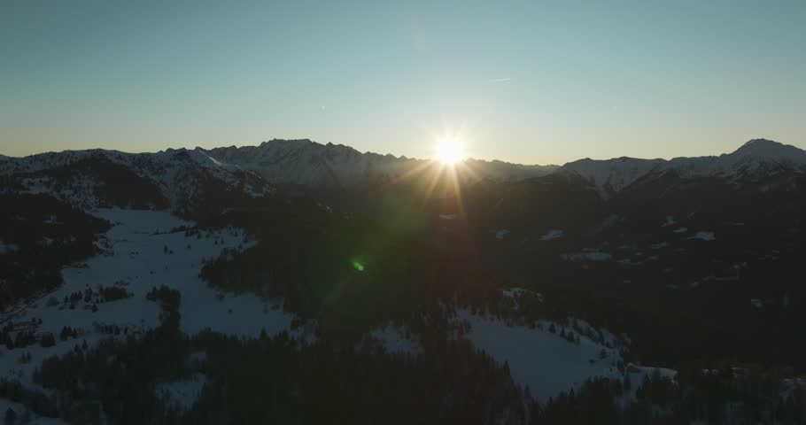 Aerial 5k sunrise over snowy alpine mountains with natural sun flare as warm rays illuminate the winter landscape, creating a cinematic and serene high-altitude scene.
