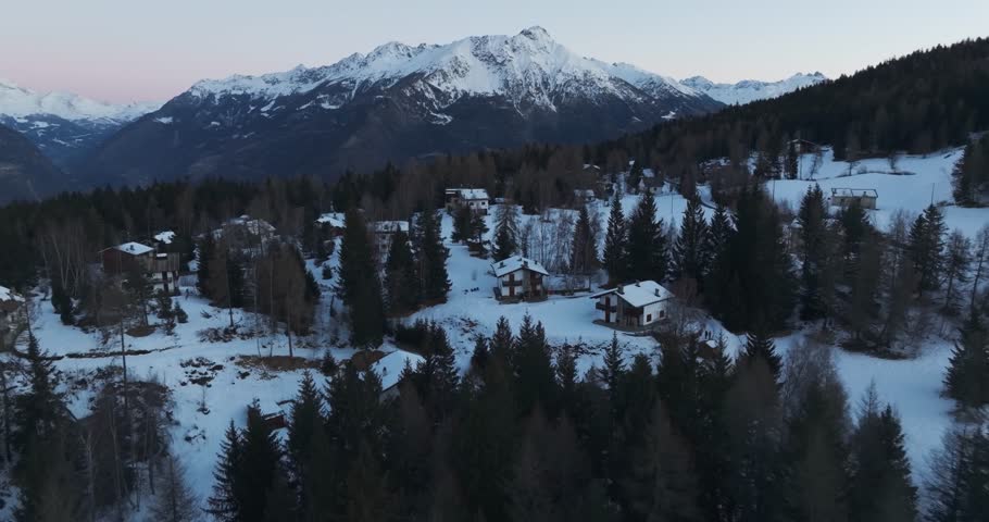 Aerial 5K winter view of a snowy alpine village surrounded by dense conifer forests and towering mountains at dusk, capturing the peaceful atmosphere of the frozen landscape.