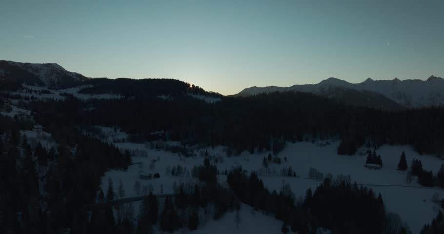 Aerial 5K sunrise over snowy alpine valleys as the sun rises behind the ridge, creating natural lens flare and illuminating the winter landscape with soft morning light.