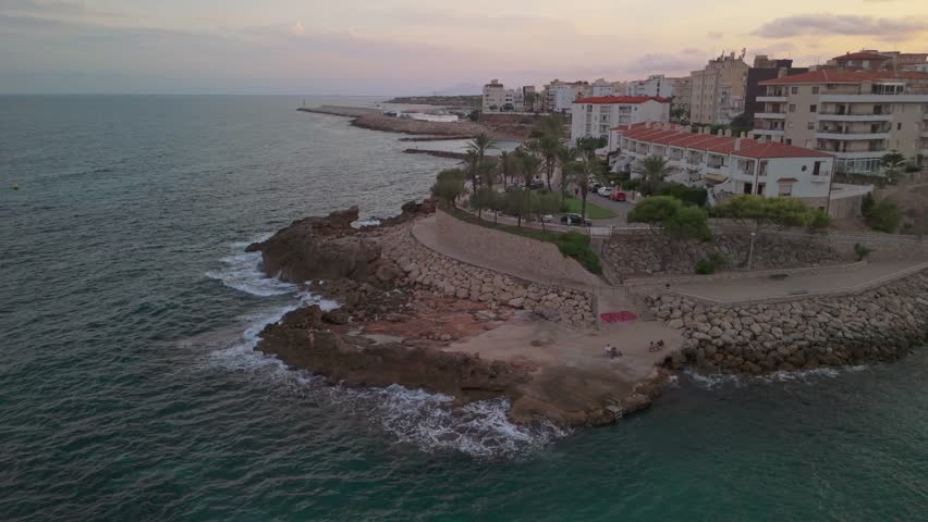 Drone orbit of rocky Mediterranean coast near L’Ametlla de Mar with waves breaking along eroded formations and white seaside houses in the background.