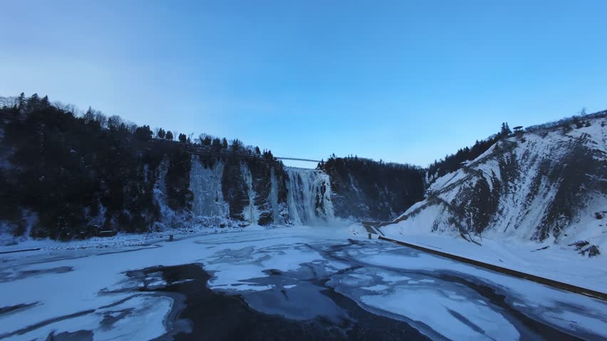 Cinematic aerial view of a frozen river and snowy waterfall in winter. Cold season nature, icy water surface and dramatic cliffs captured by drone.
