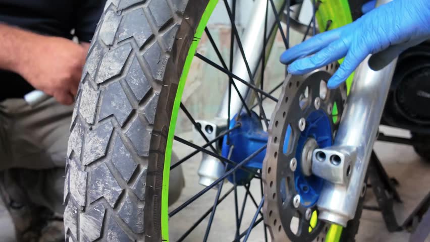 Close-up mechanics servicing motorcycle front wheel. One person holds wheel assembly while another stabilizes brake disc. Tire tread, spokes, brake rotor, and front suspension during maintenance
