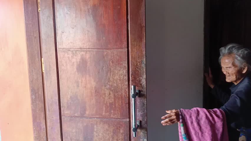 Elderly Asian woman gripping a rustic wooden door handle for stability and support as she slowly walks out of her home. Focus on aging, mobility, and the struggles of daily life in old age.