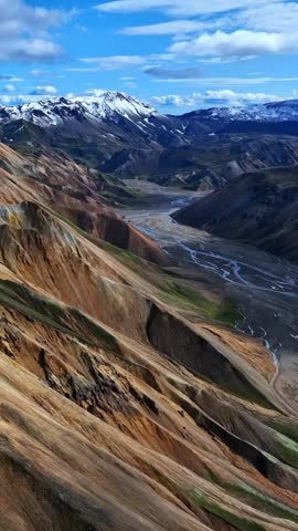 Colorful Icelandic ridges drop into a broad valley carved by braided rivers, with distant snow-covered peaks rising beneath crisp blue skies and drifting clouds.