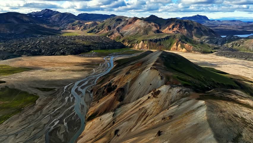 An aerial glide follows a bright rhyolite ridge overlooking a wide braided river plain, revealing eroded textures, soft summer light, and colorful volcanic layers across the Icelandic highlands.