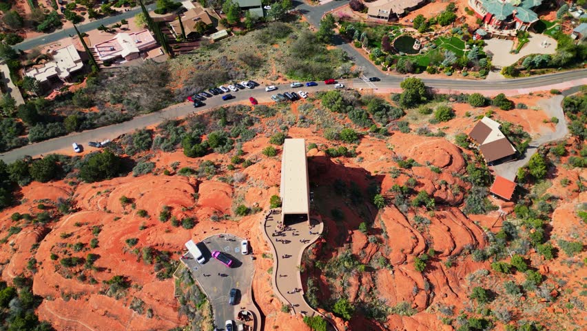 An aerial view showcases the Chapel of the Holy Cross nestled within Sedonas red rock formations, revealing residential areas and distant mountains under a bright, sunny sky.