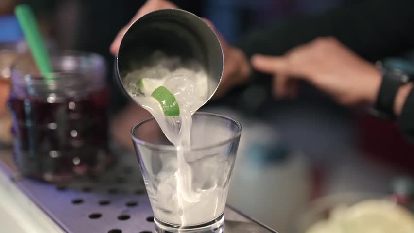 Bartender pouring a chilled lime cocktail with ice cubes into a glass at bar hall.