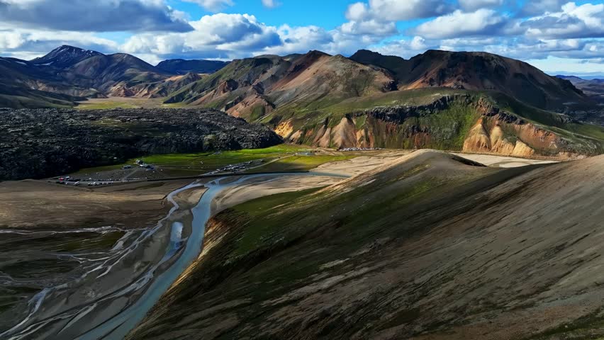 Soft sunlight washes over Iceland’s multicolored highlands as a braided river winds through a wide valley, revealing layered ridges, green plains, and volcanic textures under drifting summer clouds.