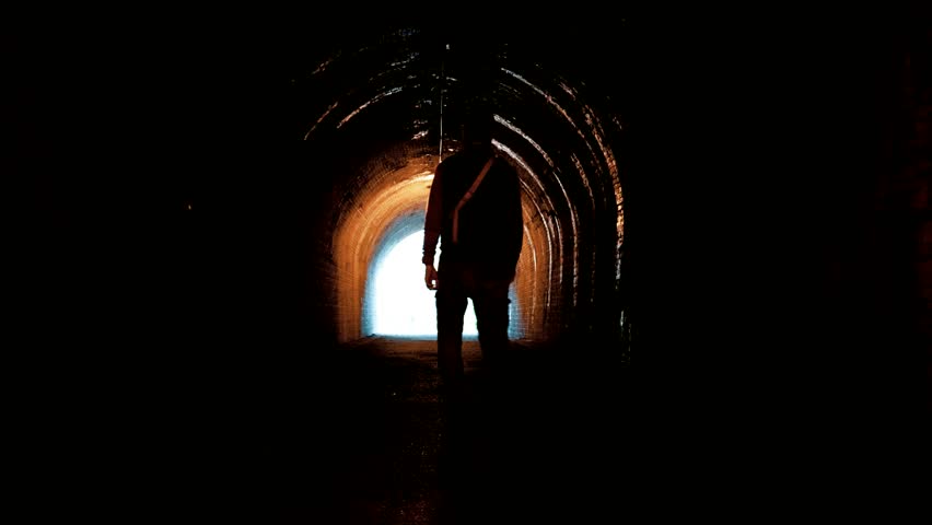 a silhouette of a man walking through a dark tunnel toward the exit.,
